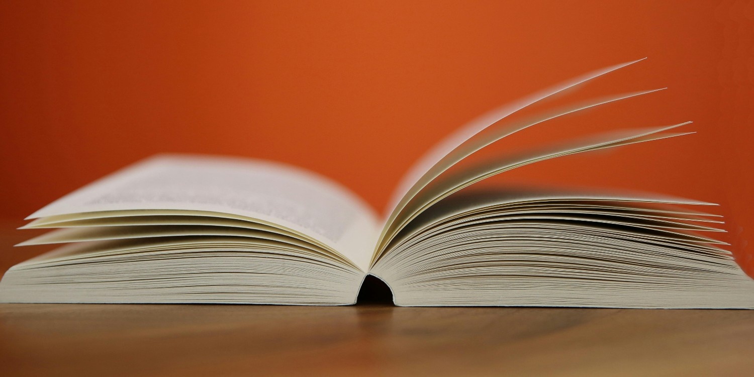 Side view of an open hardcover book with pages flipping on a wooden table against a vibrant orange background.