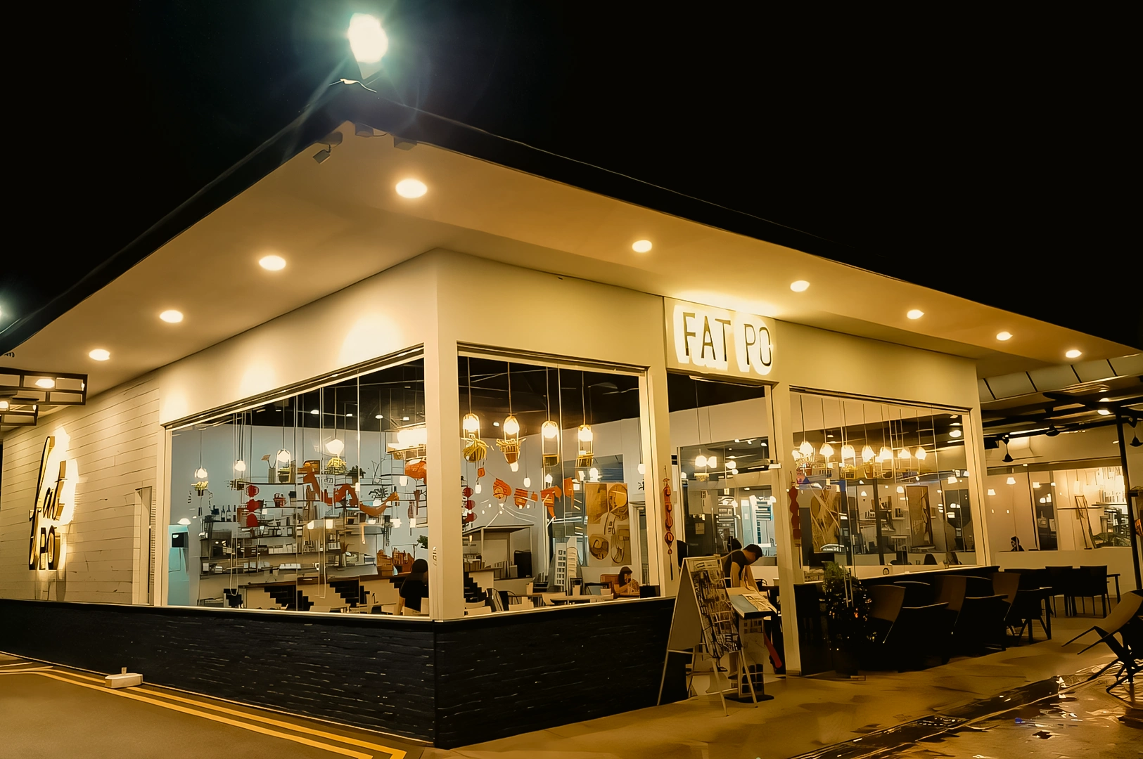 Wide-angle exterior night shot of Fat Po restaurant in Punggol with floor-to-ceiling glass windows, warm interior lighting, visible diners, and a modern white building facade.