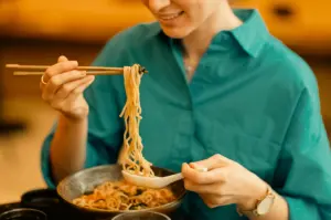 Eye-level medium close-up of a diner lifting noodles with chopsticks from a warm bowl of comfort food, capturing familiar flavors and the feeling of home in a casual dining setting.