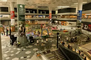 High-angle wide shot of a spacious indoor mall atrium with people walking, pop-up activity areas, and retail shops surrounding an open central space.
