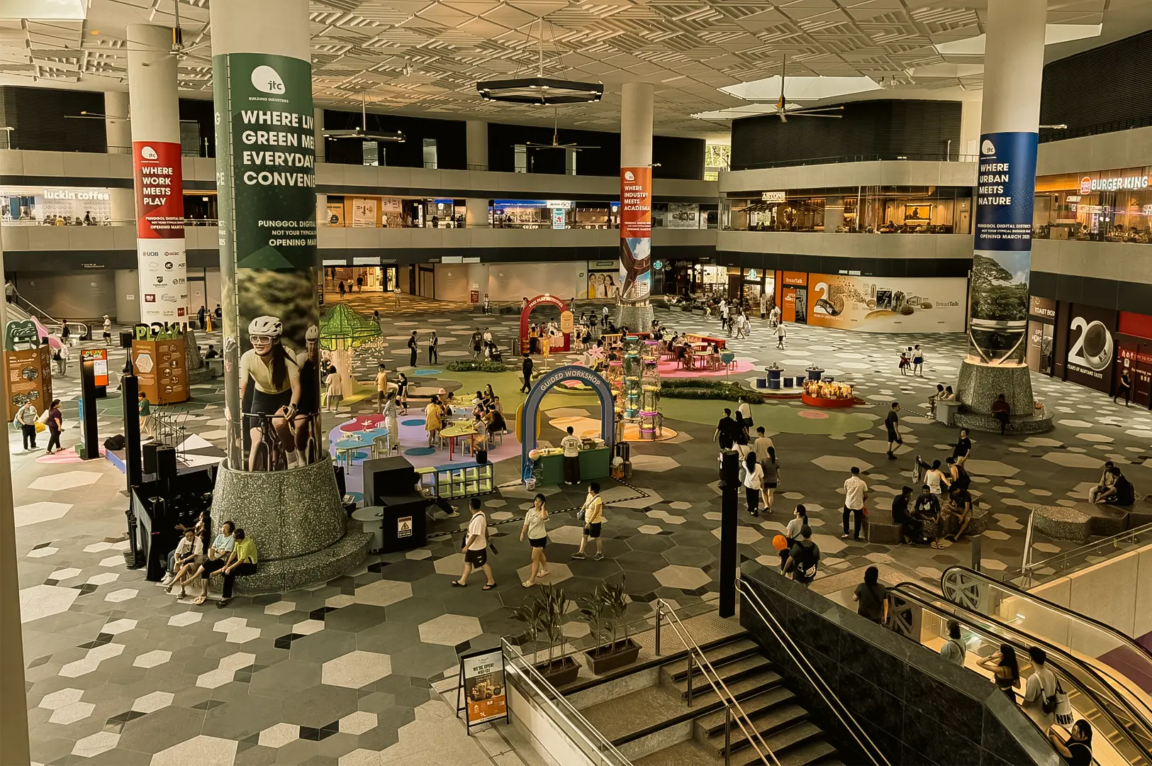 High-angle wide shot of a spacious indoor mall atrium with people walking, pop-up activity areas, and retail shops surrounding an open central space.