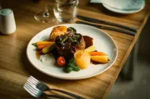 Eye-level close-up shot of a plated fine-dining dish featuring a seared meat portion with glossy sauce, root vegetables, and baby corn arranged on a ceramic plate on a wooden table, highlighting balance and presentation.
