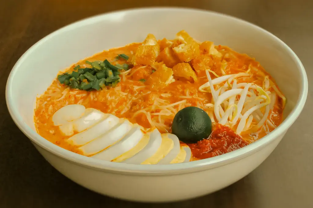 Eye-level close-up shot of a bowl of Mee Siam topped with sliced egg, bean sprouts, chives, and crispy garnish in a rich orange gravy, emphasizing balance and traditional presentation.