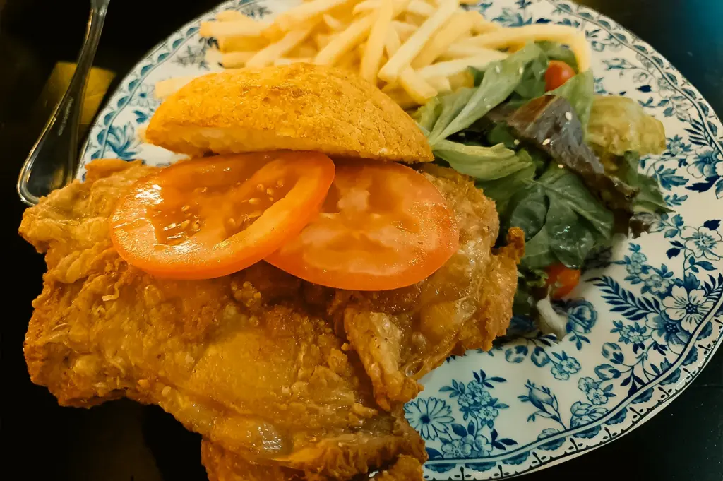 Overhead close-up shot of a crispy fried chicken burger served in a bun with tomato slices, accompanied by French fries and a side salad on a patterned ceramic plate.