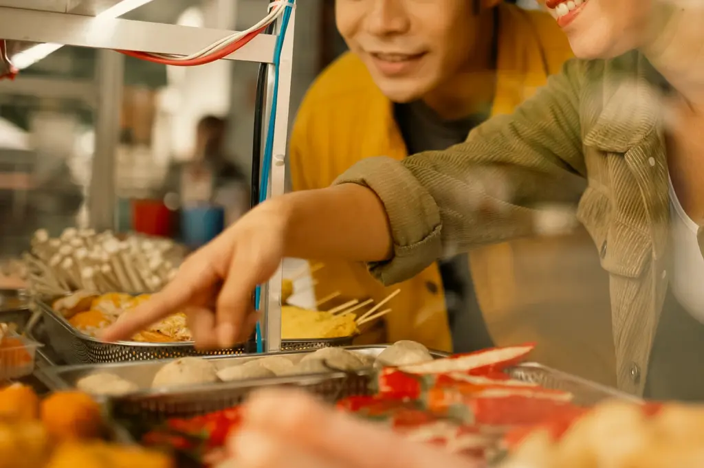 Over-the-shoulder close-up shot of diners selecting food at a casual hawker stall in Singapore, showcasing fresh ingredients and affordable local dishes in a relaxed dining atmosphere.