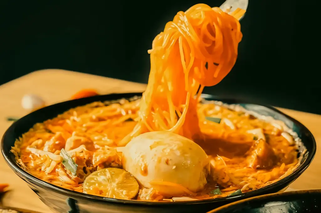 Close-up eye-level shot of Mee Siam noodles lifted from a bowl, showing orange gravy coating the noodles alongside egg and lime, highlighting texture and motion.
