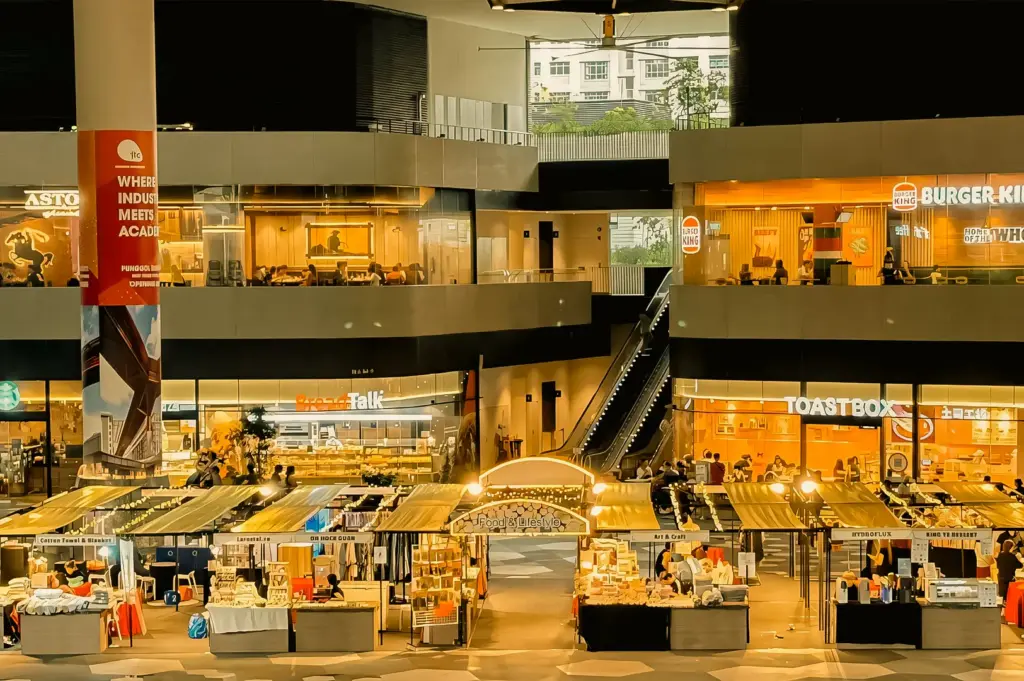 Wide-angle elevated shot of a multi-level shopping mall with illuminated cafés and food stalls on the ground floor, viewed from an upper terrace at night.
