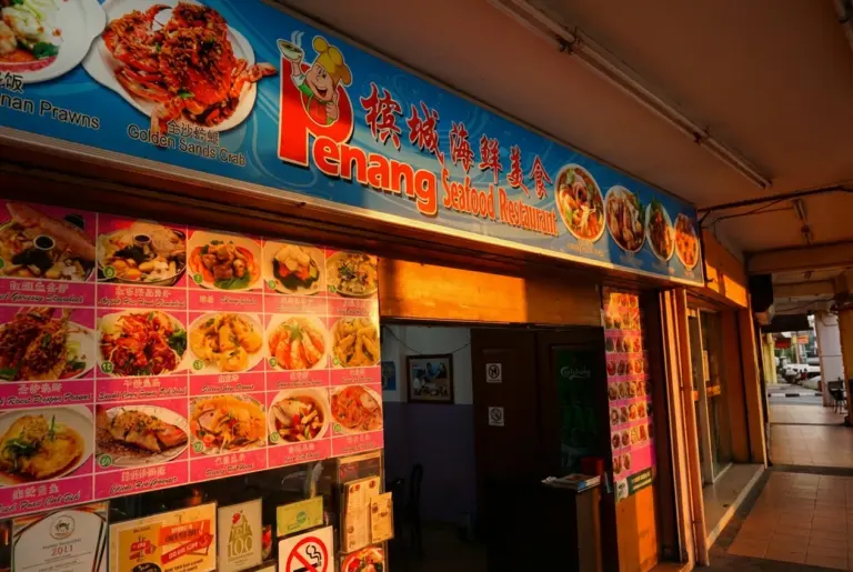 The exterior storefront of "Penang Seafood Restaurant" in Geylang, Singapore, during the golden hour. A large blue sign features colorful images of signature dishes like Golden Sands Crab and Penang Prawns. The entrance is flanked by a pink menu board displaying various zi char dishes, capturing the casual and authentic street-side dining vibe typical of the area.