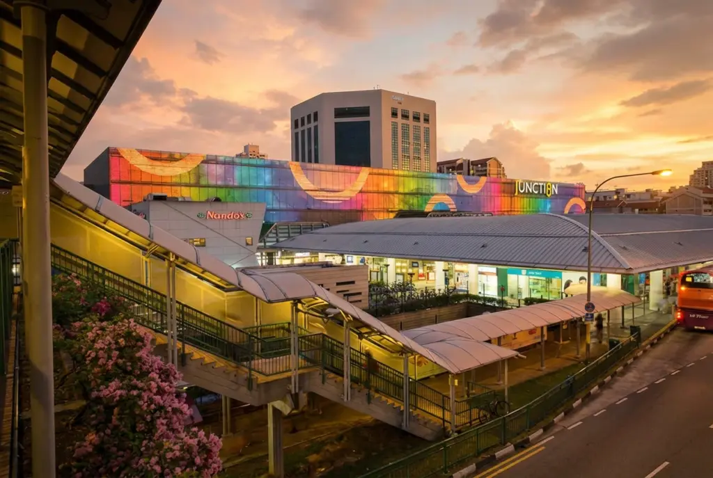 A twilight view of the Junction 8 shopping mall in Bishan, showcasing its large, colorful illuminated facade and a covered pedestrian walkway leading toward the building's entrance.