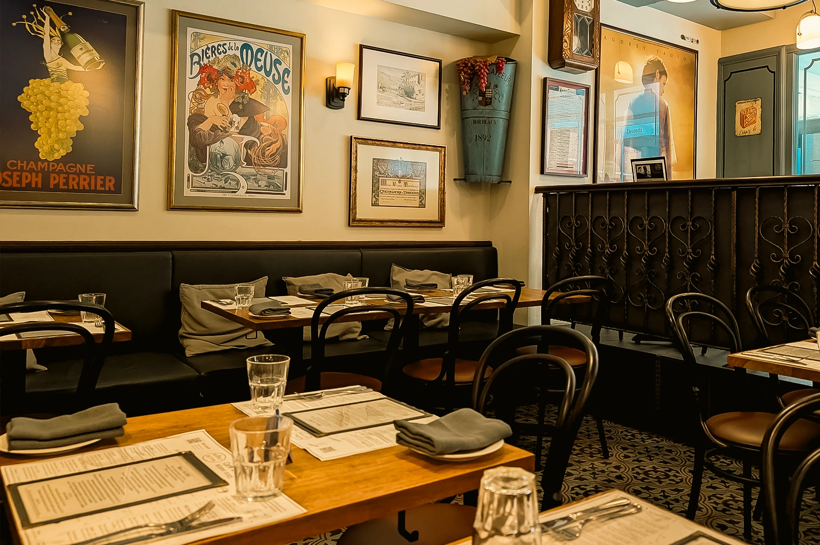 “Wide-angle eye-level view of a cozy bistro-style restaurant dining room featuring wooden tables set with menus, folded napkins, glassware, and cutlery. Black bentwood chairs and a long upholstered banquette line the seating area, while framed vintage-style posters, artwork, and a wall-mounted clock decorate the cream-colored walls. Ornate black railing details, patterned floor tiles, and warm ambient lighting create an inviting and classic dining atmosphere with a refined yet casual interior design.”