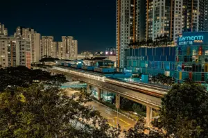 Night cityscape of Clementi MRT station and The Clementi Mall in Singapore, featuring elevated train tracks with light trails, surrounded by high-rise residential buildings and urban greenery, captured from a high-angle wide shot