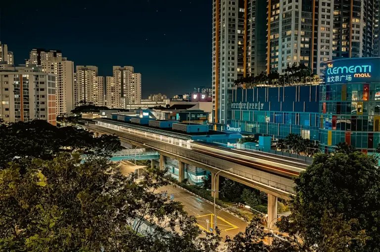 Night cityscape of Clementi MRT station and The Clementi Mall in Singapore, featuring elevated train tracks with light trails, surrounded by high-rise residential buildings and urban greenery, captured from a high-angle wide shot