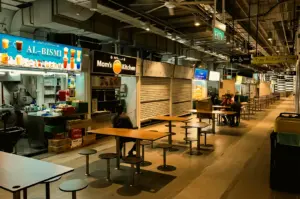 Wide-angle eye-level shot of a nearly empty indoor hawker centre at night, featuring rows of clean communal tables and stools, a few scattered diners, partially closed food stalls, warm overhead lighting, and a quiet late-evening dining atmosphere.