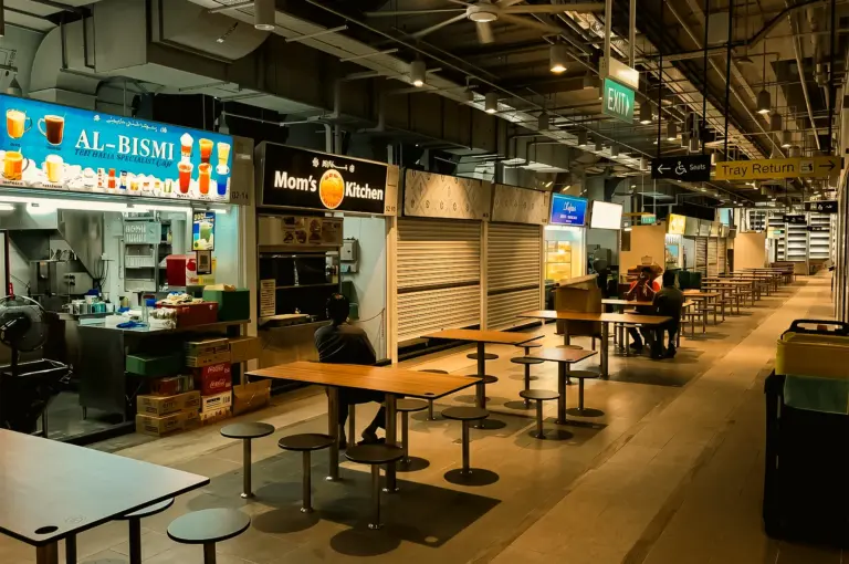 Wide-angle eye-level shot of a nearly empty indoor hawker centre at night, featuring rows of clean communal tables and stools, a few scattered diners, partially closed food stalls, warm overhead lighting, and a quiet late-evening dining atmosphere.