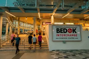 Wide-angle street-level shot of visitors entering Bedok Interchange Hawker Centre beside Bedok MRT, showcasing a busy Singapore neighbourhood dining hub with local food stalls.