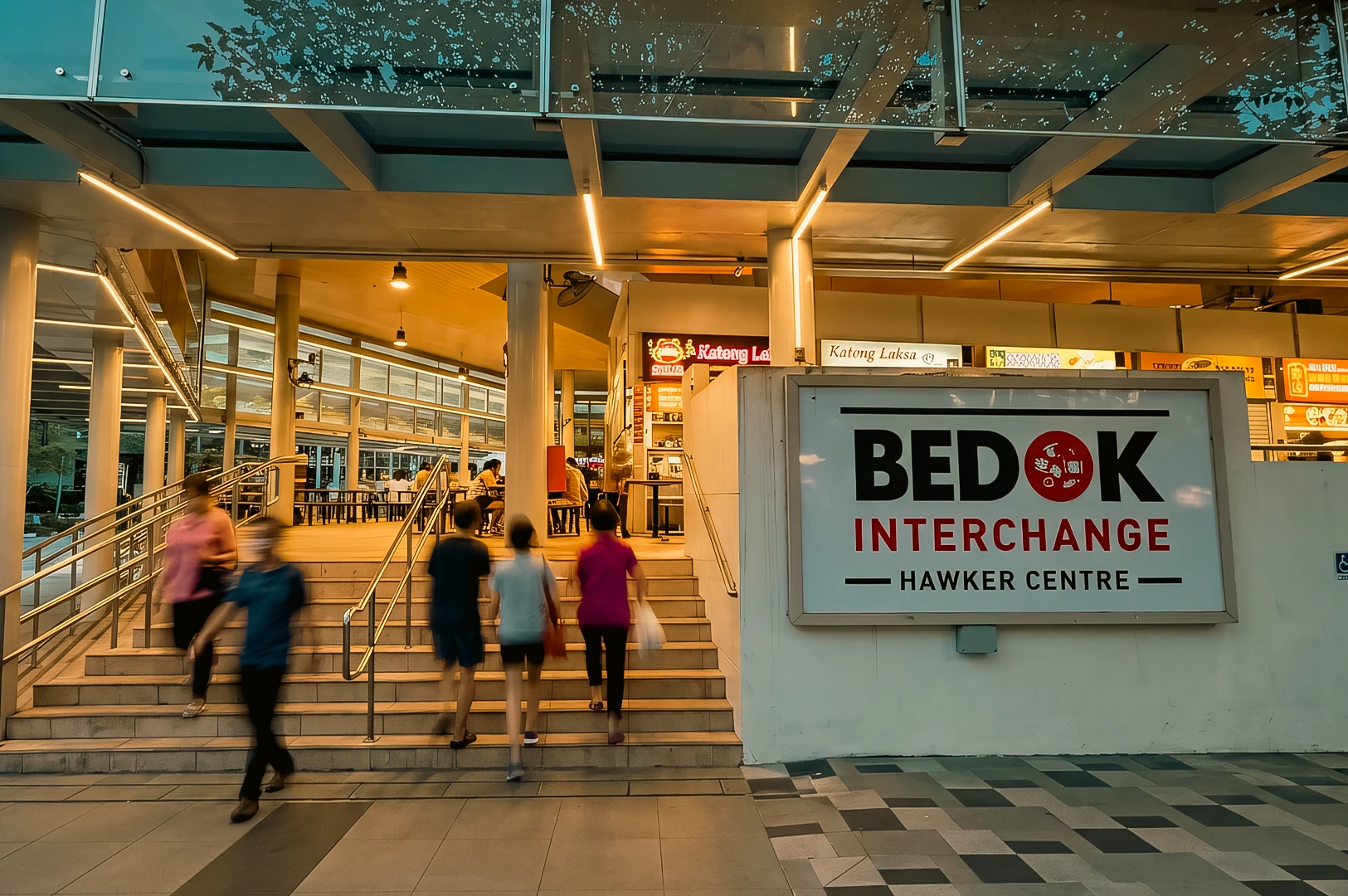 Wide-angle street-level shot of visitors entering Bedok Interchange Hawker Centre beside Bedok MRT, showcasing a busy Singapore neighbourhood dining hub with local food stalls.