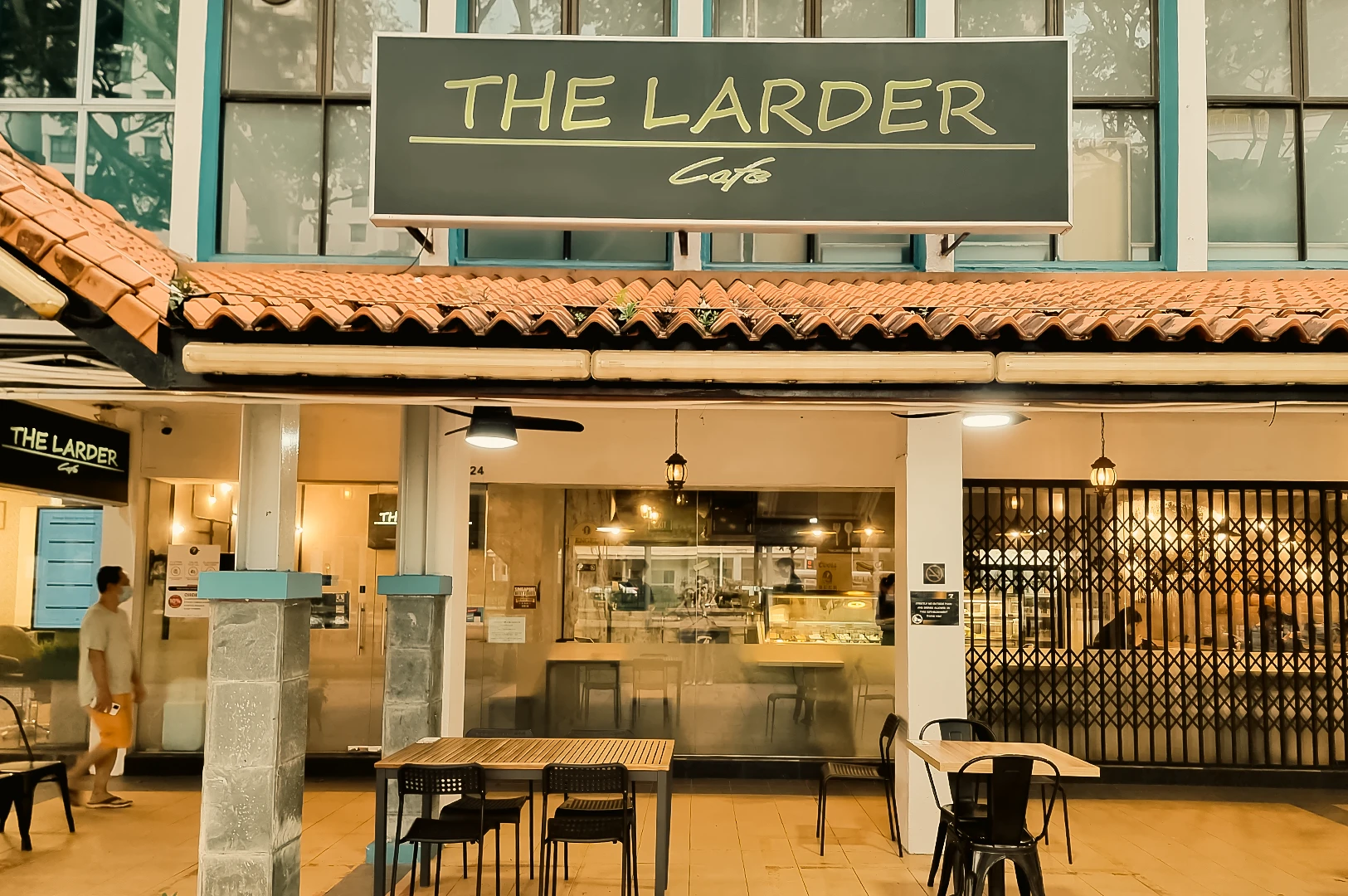 The Larder Cafe exterior in Toa Payoh with rustic tiled roof, warm lighting, and outdoor seating area, captured in a straight-on eye-level shot highlighting the storefront signage and inviting facade.