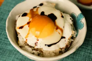 Close-up, slightly overhead shot of a sunny-side-up egg with a glossy, runny yolk drizzled in soy sauce over a bowl of steamed white rice, served in a small ceramic bowl as simple Japanese comfort food.