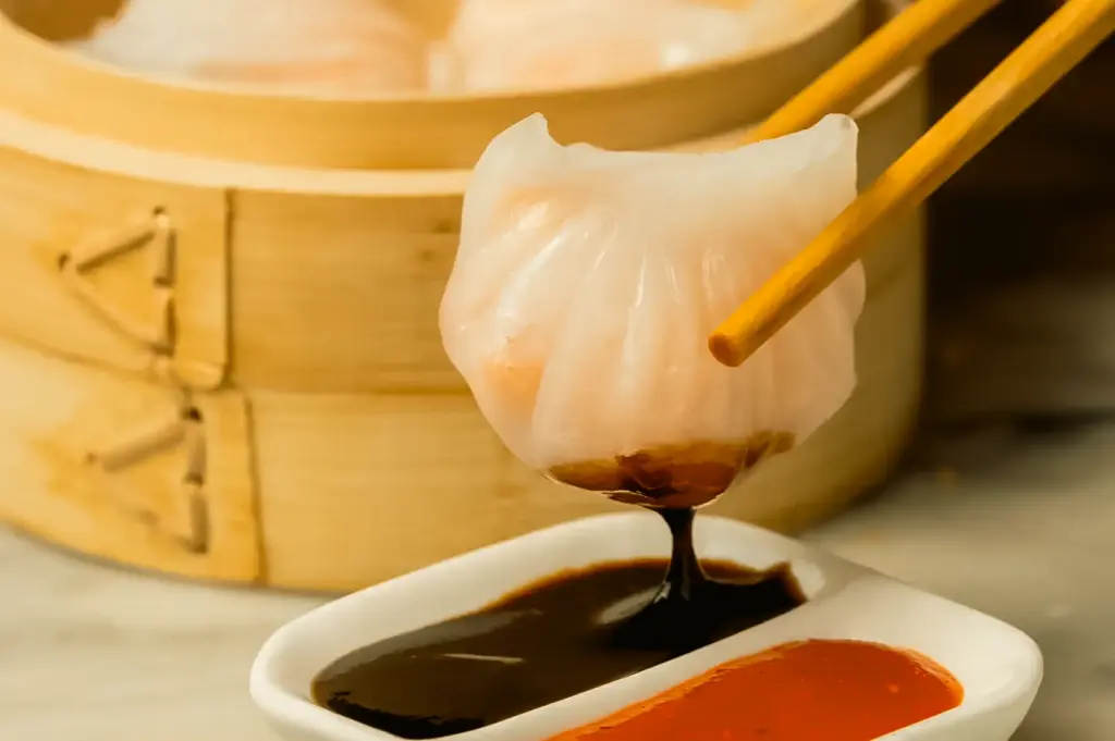 Close-up macro shot of steamed shrimp dim sum dumpling (har gow) held with chopsticks dipping into soy sauce and chili oil, bamboo steamer in background, traditional dim sum Singapore lunch