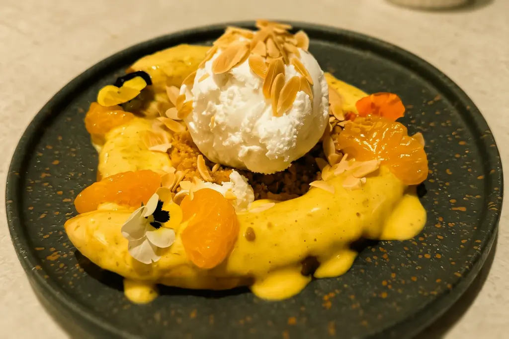 Close-up slightly top-down shot of Banana Orange Foster dessert at Clovelly Serangoon featuring coconut gelato, mandarin orange slices, toasted almonds, and creamy sabayon sauce