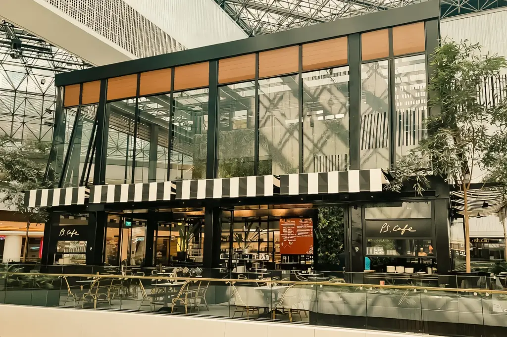 Contemporary café exterior with glass facade inside a mall atrium, lit by natural daylight filtering through the roof mixed with soft interior lighting, captured in a wide-angle exterior shot.