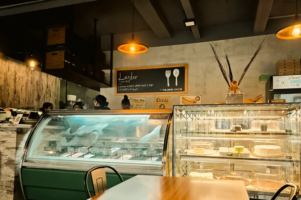The Larder Cafe dessert and gelato display counter with cakes, chalkboard menu, and warm ambient lighting, captured in a slightly low-angle interior shot highlighting the dessert selection and cozy cafe atmosphere.