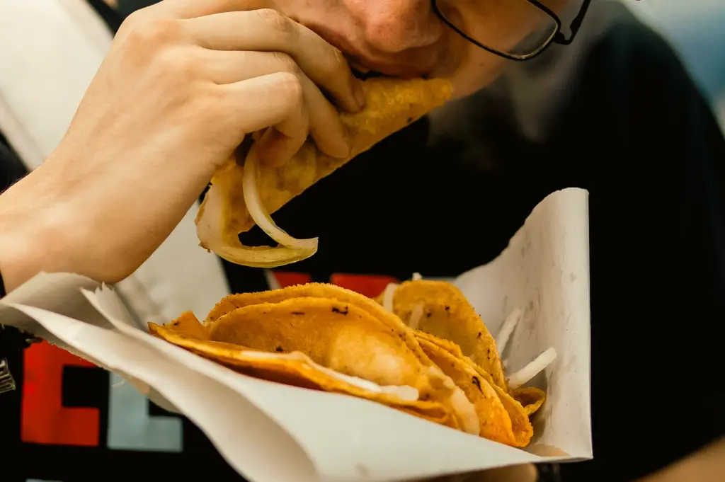 Close-up candid shot of person eating crispy street tacos from takeaway tray, portable fast lunch street food experience, taco truck style dining