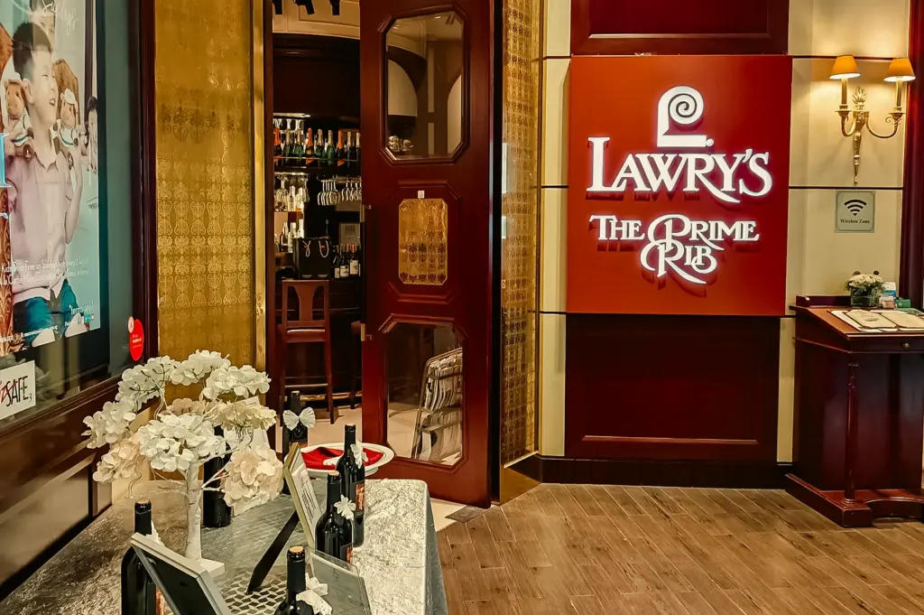 Lawry’s The Prime Rib restaurant entrance with red signage and polished wood interiors, highlighted by warm accent lighting and soft overhead illumination, captured in a frontal mid-range shot.