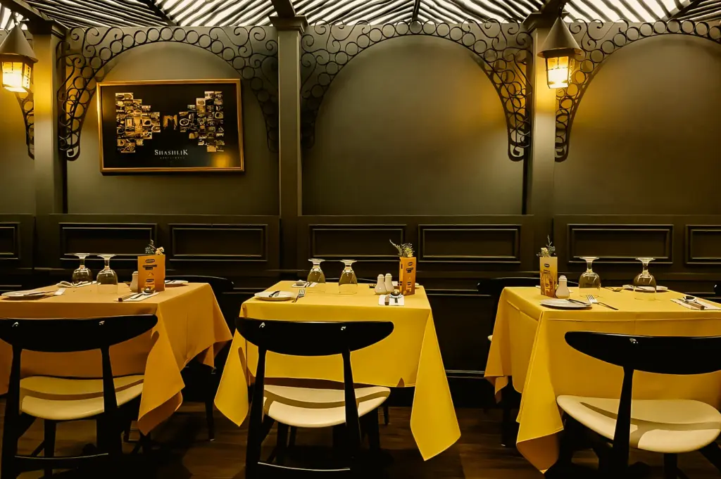 Upscale restaurant interior with neatly arranged tables and yellow tablecloths under decorative arches, lit by warm wall sconces and diffused ceiling lighting, captured in a straight-on wide-angle shot.