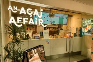 An eye-level, wide-angle exterior shot of the glass storefront of An Açaí Affair at Robertson Quay, showcasing the bright white illuminated logo on the window, indoor service counter, wall menus, and a green potted plant framing the entrance.
