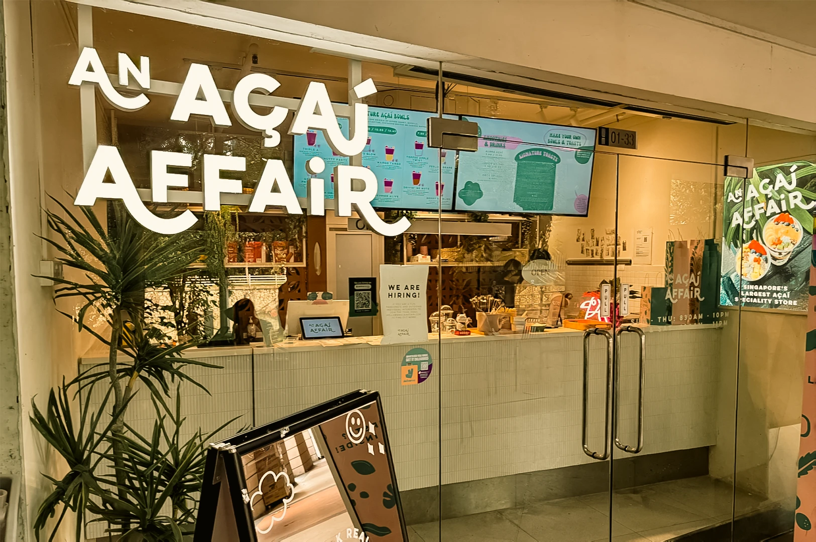 An eye-level, wide-angle exterior shot of the glass storefront of An Açaí Affair at Robertson Quay, showcasing the bright white illuminated logo on the window, indoor service counter, wall menus, and a green potted plant framing the entrance.