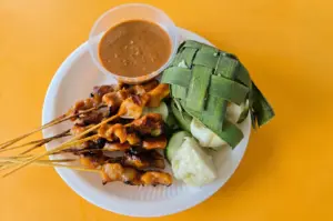 Top-down shot of Singapore satay skewers with peanut sauce, ketupat rice cakes wrapped in palm leaves, and cucumber slices on a white plate against a bright yellow table, highlighting familiar hawker comfort food.