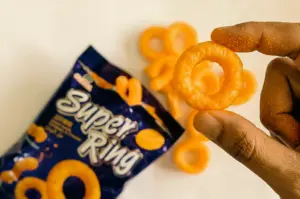 Close-up macro shot of a hand holding a Super Ring cheese snack, highlighting the orange ring-shaped texture with blurred packaging in the background, nostalgic Singaporean snack