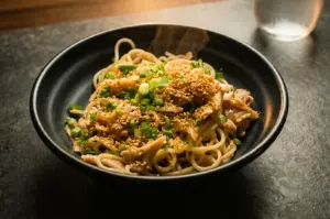 A slightly high-angle, three-quarter close-up shot of a steaming bowl of fusion pasta or noodles served in a matte black ceramic bowl, topped with thin slices of pork, chopped green onions, and a generous sprinkling of toasted sesame seeds, set against a dark textured tabletop with a glass of water softly blurred in the background.