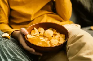 Close-up, slightly top-down shot of a person holding a wooden bowl filled with crispy potato chips on a couch, cozy at-home snacking scene with warm lighting ideal for comfort food and snack lifestyle SEO.