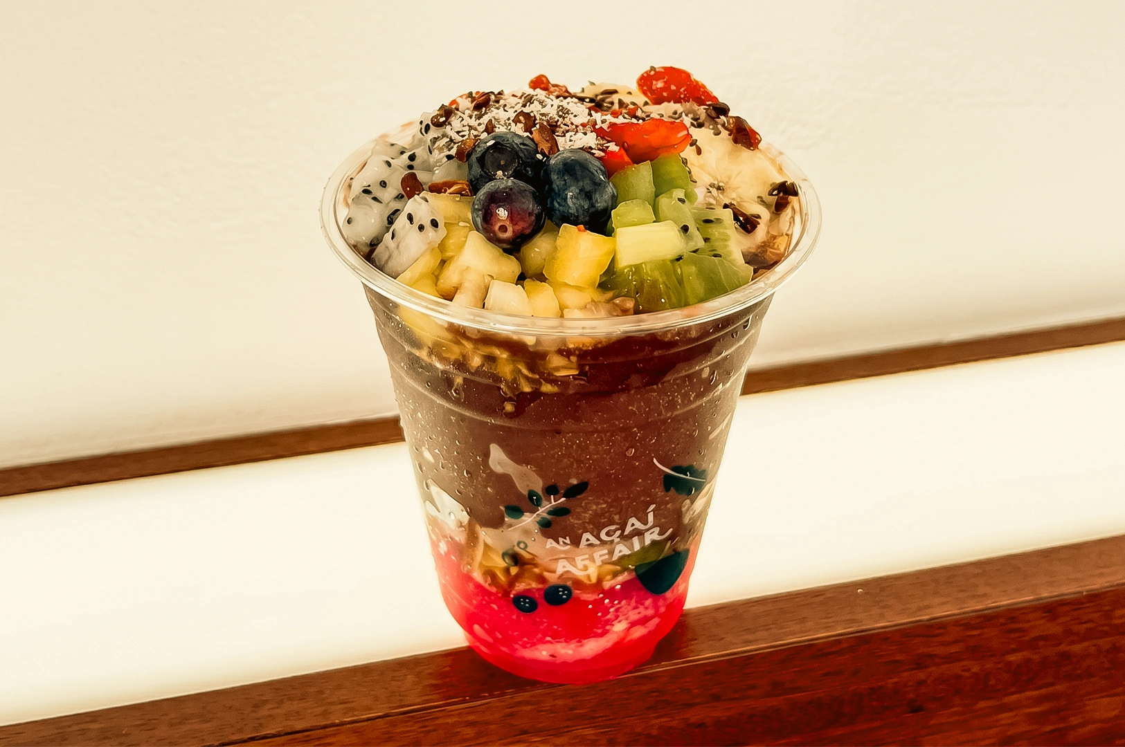 Close-up slightly top-down shot of a fresh acai bowl in a clear cup topped with strawberries, blueberries, kiwi, dragon fruit, granola, and coconut flakes, perfect healthy snack before heading home