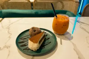 Eye-level close-up of a creamy tiramisu slice with chocolate drizzle and a scoop of chocolate ice cream on a plate, paired with a glass of iced coffee on a marble table in a cozy cafe interior, highlighting a calm and indulgent dessert moment.