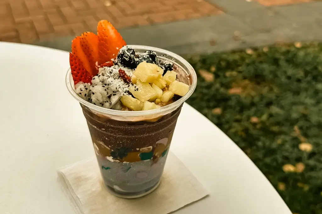 A slightly high-angle, medium close-up shot of a signature Triple A açaí bowl from An Açaí Affair, served in a tall clear plastic cup on an outdoor white table, featuring neatly layered sliced strawberries, diced dragonfruit, pineapple chunks, blueberries, and coconut shavings over a dark açaí base.