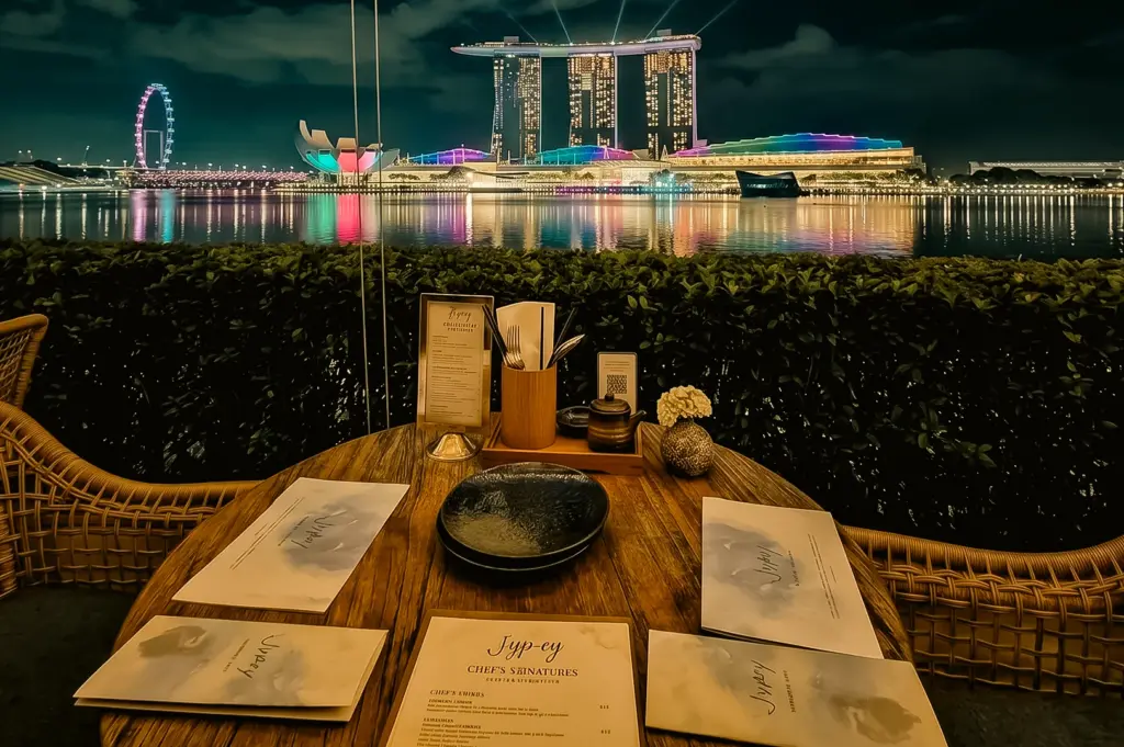 Eye-level night shot of a romantic table setup at Jypsy One Fullerton overlooking Marina Bay Sands skyline, highlighting waterfront dining Singapore with city lights and intimate date night atmosphere.