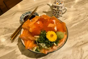 “Three-quarter, close-up view of a salmon ikura don served in a ceramic bowl on a marble table, featuring thick slices of fresh salmon sashimi arranged around a generous mound of salmon roe, chopped green onions, a yellow edible flower garnish, and a scoop of wasabi over rice. Gold chopsticks rest beside a small dish of soy sauce, while a patterned ceramic teapot sits in the background. Warm indoor lighting highlights the glossy seafood textures, vibrant orange tones, and elegant Japanese dining presentation.