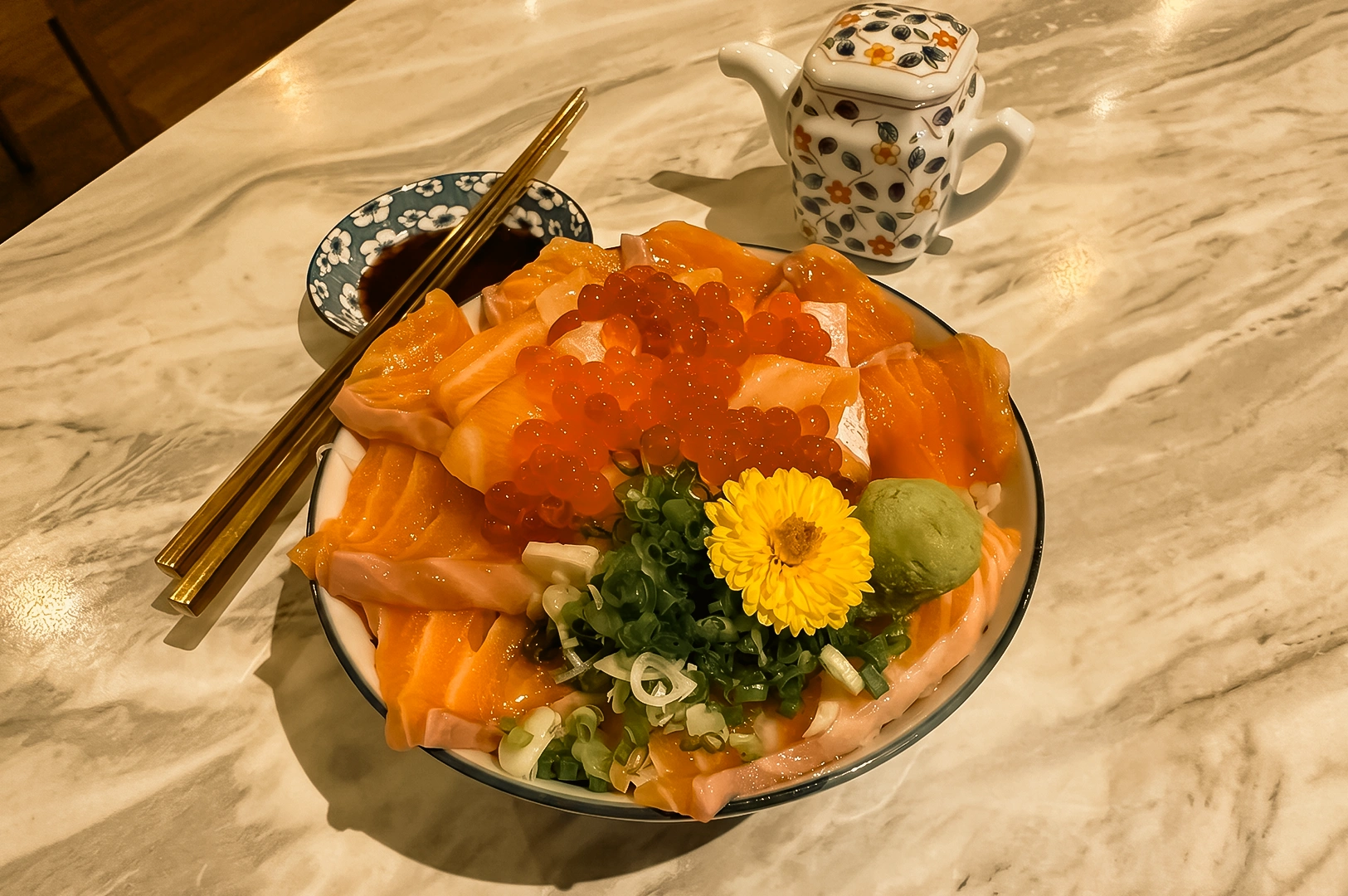 “Three-quarter, close-up view of a salmon ikura don served in a ceramic bowl on a marble table, featuring thick slices of fresh salmon sashimi arranged around a generous mound of salmon roe, chopped green onions, a yellow edible flower garnish, and a scoop of wasabi over rice. Gold chopsticks rest beside a small dish of soy sauce, while a patterned ceramic teapot sits in the background. Warm indoor lighting highlights the glossy seafood textures, vibrant orange tones, and elegant Japanese dining presentation.