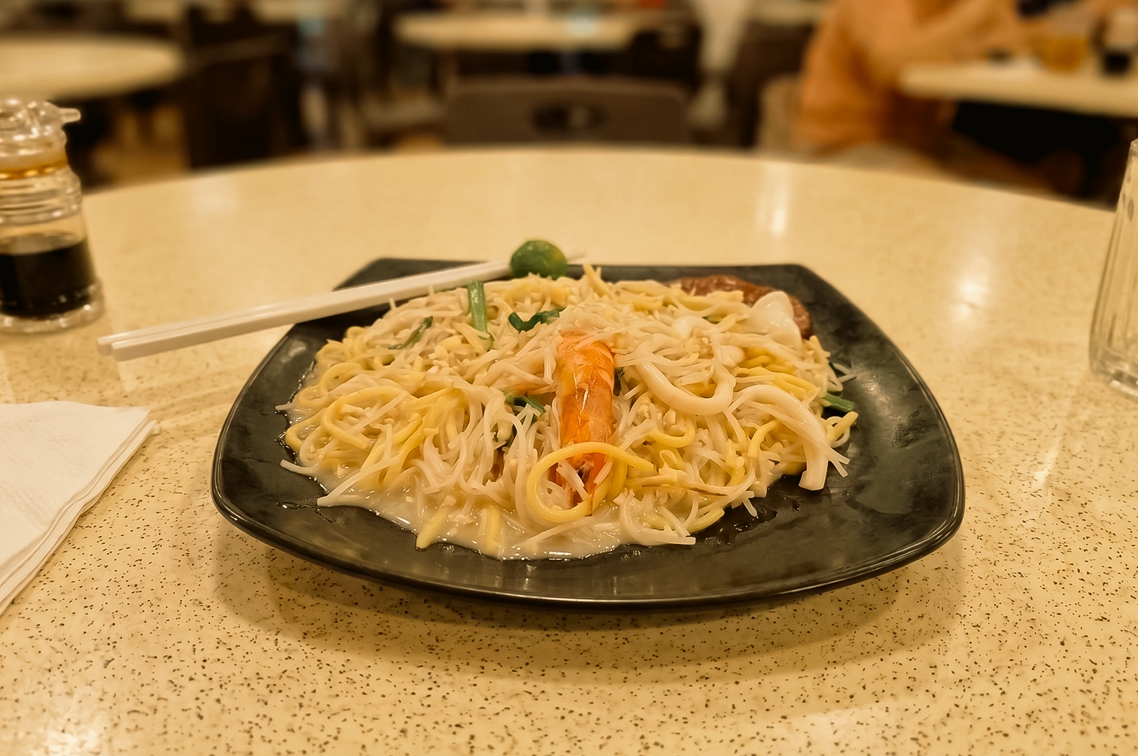 Eye-level close-up of a plate of traditional Hokkien Mee at a Pasir Ris coffeeshop in Singapore, featuring springy yellow noodles, rice vermicelli, prawns, squid, bean sprouts, and calamansi in a classic neighbourhood dining setting.