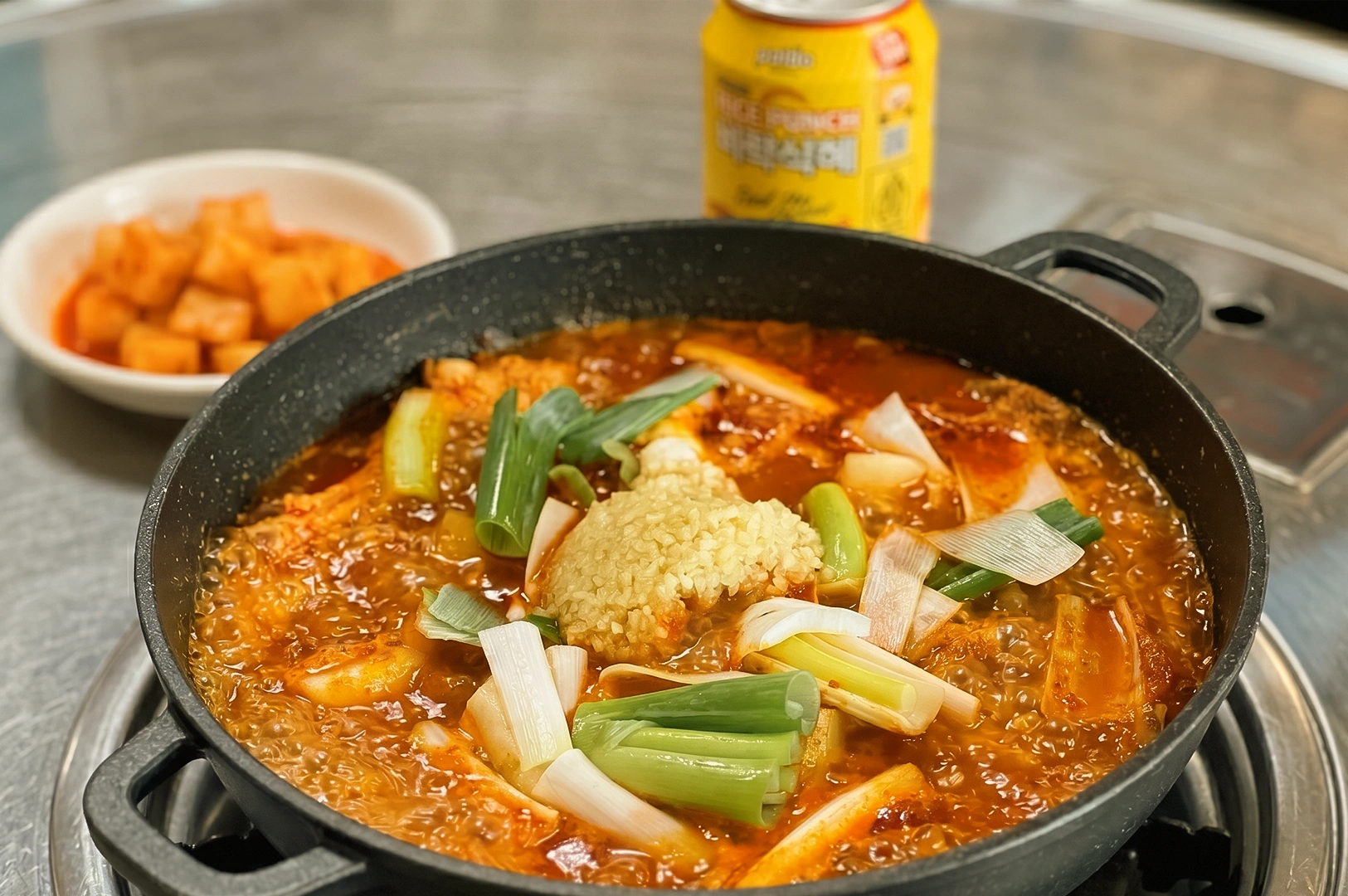 Close-up eye-level shot of a bubbling Korean dakdoritang stew in a black pot with rice, green onions, and rich spicy broth on a tabletop stove, accompanied by cubed radish kimchi and a soda can for an authentic solo dining experience.