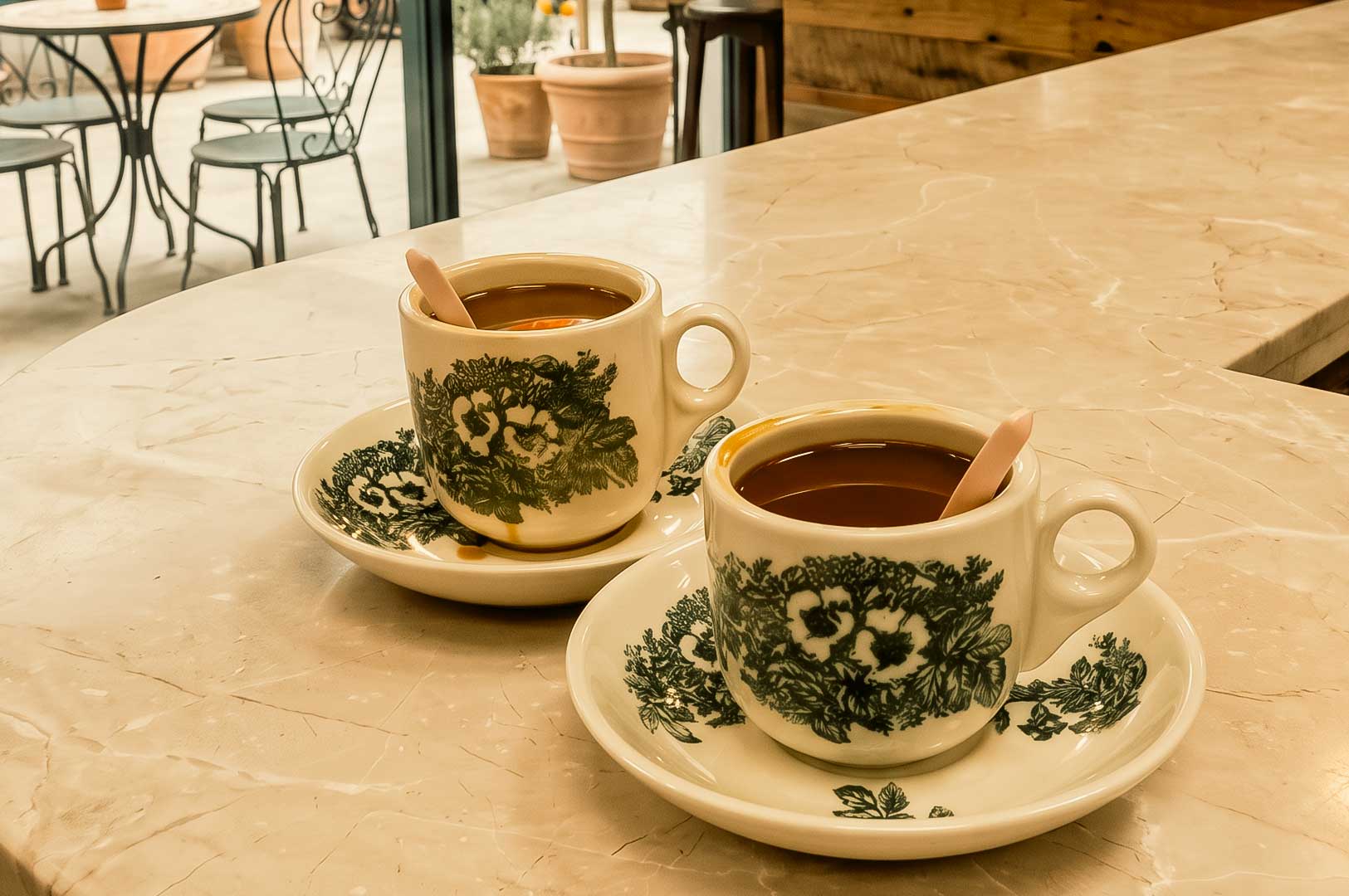 Two ceramic cups of hot coffee sit on matching saucers atop a marble countertop, each with a spoon resting inside. In the background, an outdoor patio with metal café tables, chairs, and potted plants is softly lit by natural daylight, creating a warm and inviting coffee shop atmosphere.