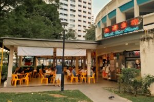 Alt text: Exterior view of a casual open-air restaurant with a bright neon sign reading "Cheah Dee's Hainanese Village." Yellow plastic tables and chairs are filled with diners enjoying meals, while apartment buildings and lush trees surround the eatery. A pedestrian walkway and a pigeon are visible in the foreground, creating a lively neighborhood atmosphere.