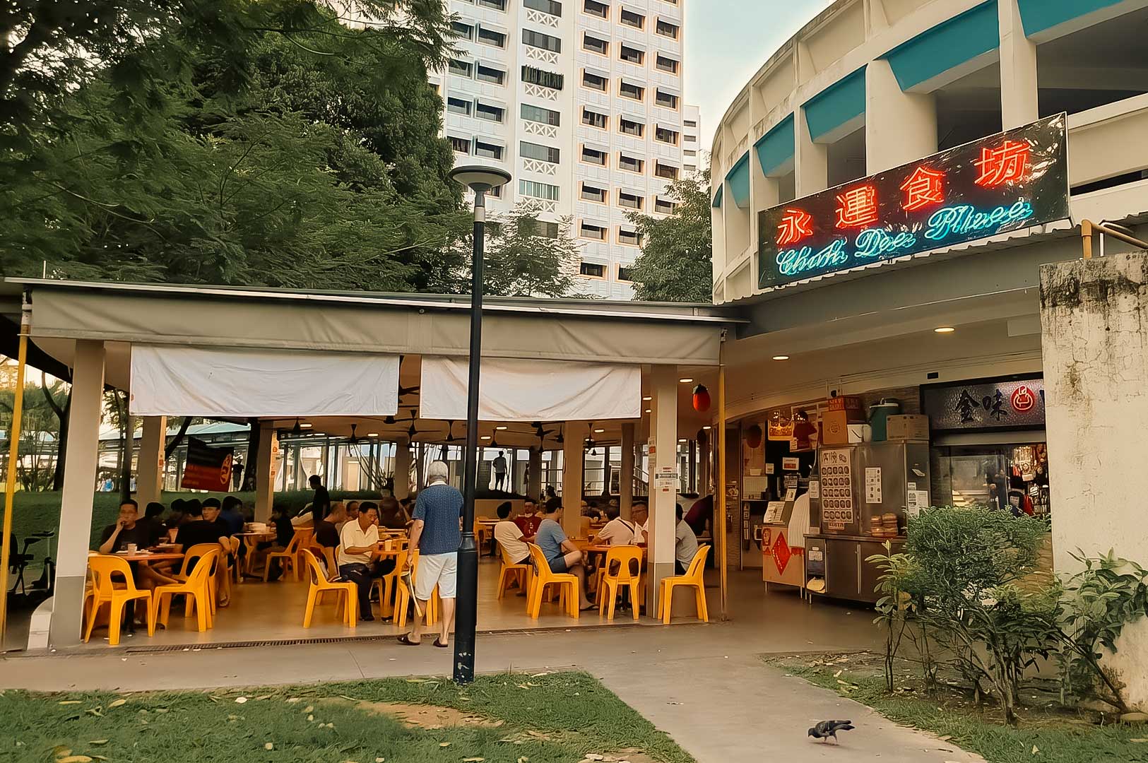 Alt text: Exterior view of a casual open-air restaurant with a bright neon sign reading "Cheah Dee's Hainanese Village." Yellow plastic tables and chairs are filled with diners enjoying meals, while apartment buildings and lush trees surround the eatery. A pedestrian walkway and a pigeon are visible in the foreground, creating a lively neighborhood atmosphere.