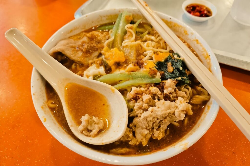 Alt text: Close-up of a steaming bowl of noodle soup on an orange table. The bowl contains curly noodles in a rich brown broth, topped with minced pork, leafy greens, dumplings, tofu, and vegetables. A white ceramic soup spoon rests inside the bowl, with chopsticks placed across the rim. A small dish of sliced chili in soy sauce sits in the background beside a tray.