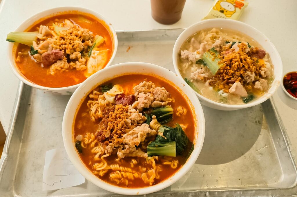 Alt Text: Three bowls of Ke Kou Mian (instant noodle soup) served on a metal tray. Two bowls feature a vibrant, reddish broth, while the third bowl has a lighter, clear broth. All three bowls are generously topped with minced meat, leafy green vegetables, and crispy fried garlic bits, with a small saucer of sliced chili padi on the side.