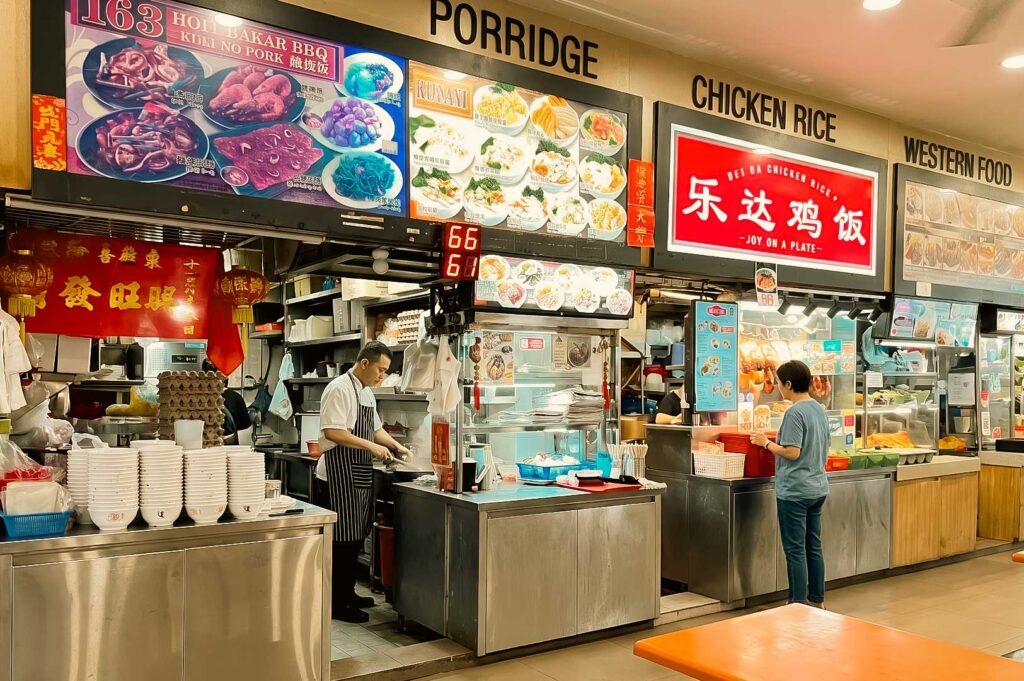 The images depict a food stall setup inside a hawker center or food court, featuring various vendors including Hoi Bakar BBQ, Kunani, and Dei Da Chicken Rice. At one of the stalls, a vendor in an apron prepares meals next to stacks of white bowls while a digital queue board shows the number 66. Customers can enjoy dishes such as Ke Kou Mian, which is served in bowls featuring either a vibrant reddish broth or a clear broth, generously topped with minced meat, leafy greens, crispy fried garlic bits, and a side of sliced chili padi.