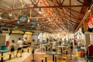 Wide view inside the bustling Maxwell Food Centre hawker hall under a high, exposed orange steel roof. Rows of food stalls line both sides of a central seating aisle filled with round green dining tables and bright yellow stools, with people motion-blurred as they walk through the space.